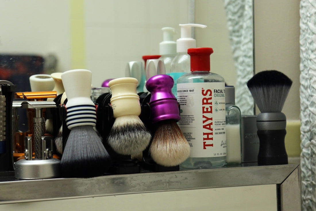 Multiple shaving brushes and skincare products on a bathroom shelf.